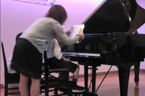 グランドピアノに座る小さな男の子の前に、開いた楽譜を置こうとしている女性