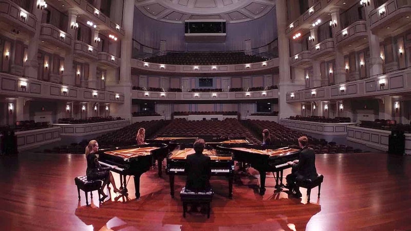 観客がいない広いホールのステージに同心円に配置された5台のグランドピアノとピアニストの写真