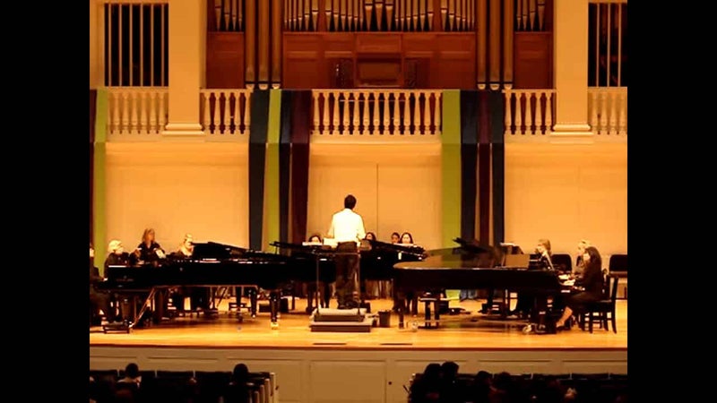 8台のグランドピアノが置かれ、中央に指揮者が立つステージを客席から写した写真