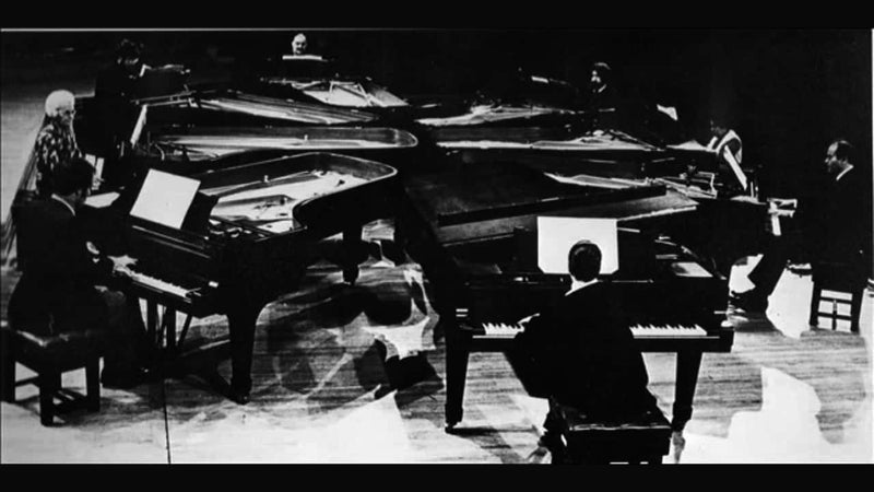同心円状に配置された8台のグランドピアノとピアニストたちの白黒の写真