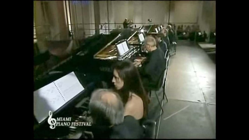 無機質なデザインのステージに、指揮者のいる奥に向かって横並びに配置された何台もののピアノを連弾している演奏家たちの写真