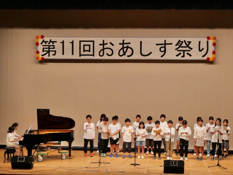 壇上に並んで合唱をする子供たちとピアノ演奏をする子供たち
